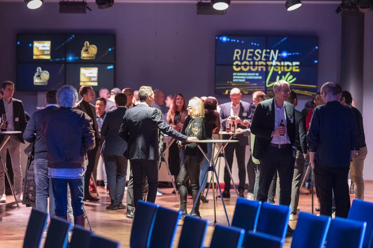 Gäste im Gespräch an Stehtischen vor der Leinwand „RIESEN Courtside Live“
